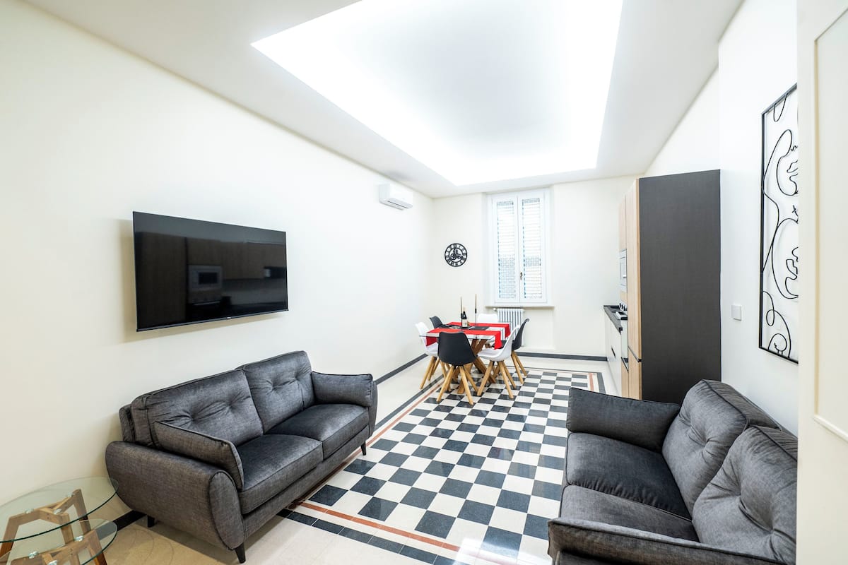 Una sala de estar moderna con dos sofás grises, una mesa de centro de vidrio, un televisor de pantalla plana montado en la pared, un comedor con una mesa y sillas rojas, y un piso a cuadros.