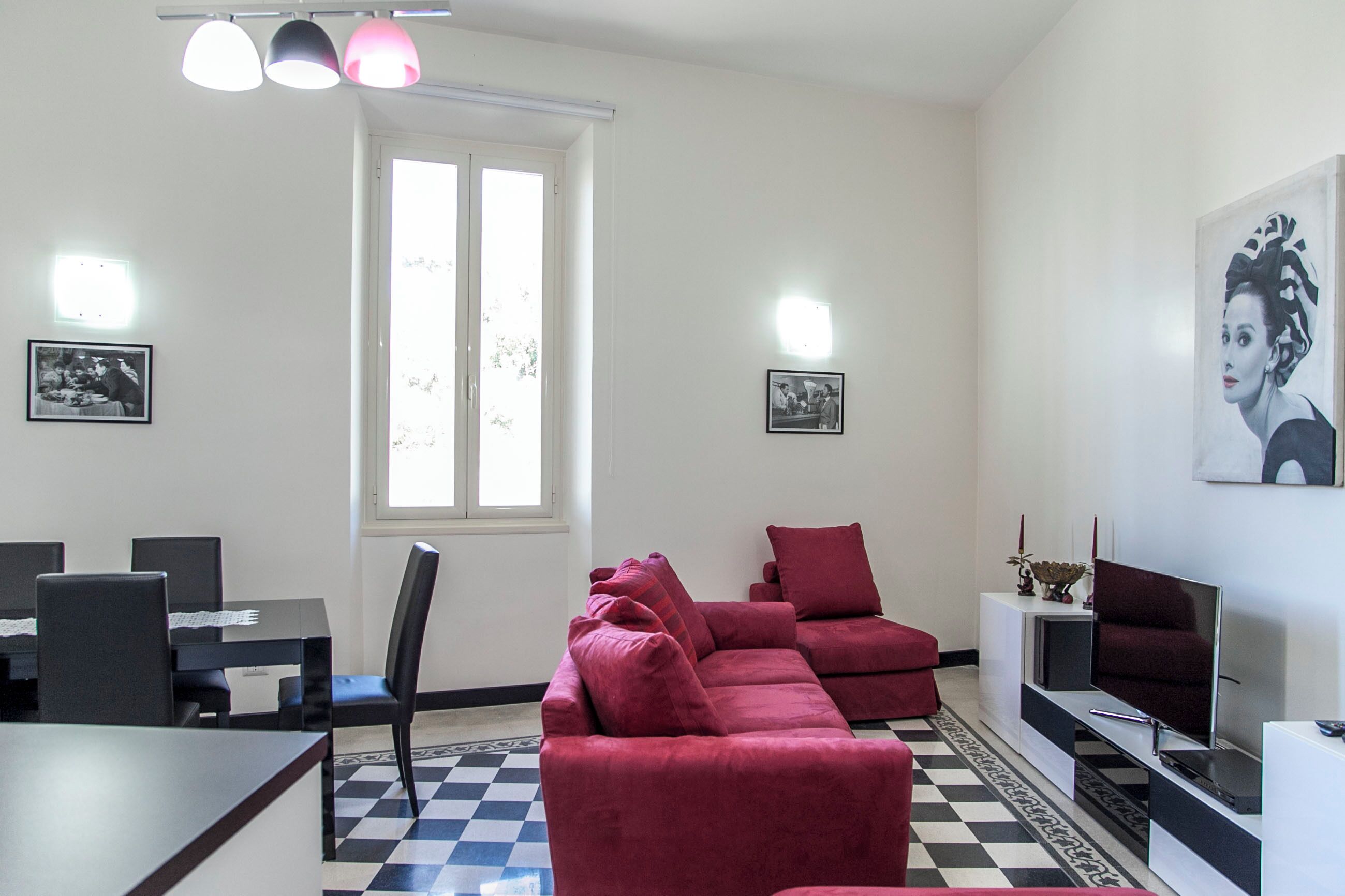 Una sala de estar con piso a cuadros, un sofá rojo, una mesa de comedor con sillas y un televisor sobre un mueble.