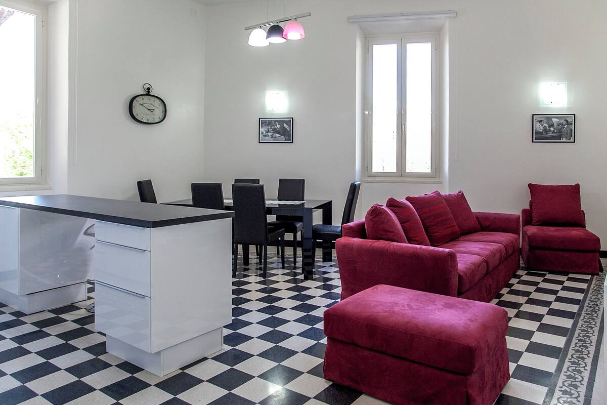 Una habitación con piso a cuadros, una mesa de comedor con sillas, un sofá rojo, un sillón rojo, una pequeña cocina y un reloj en la pared.