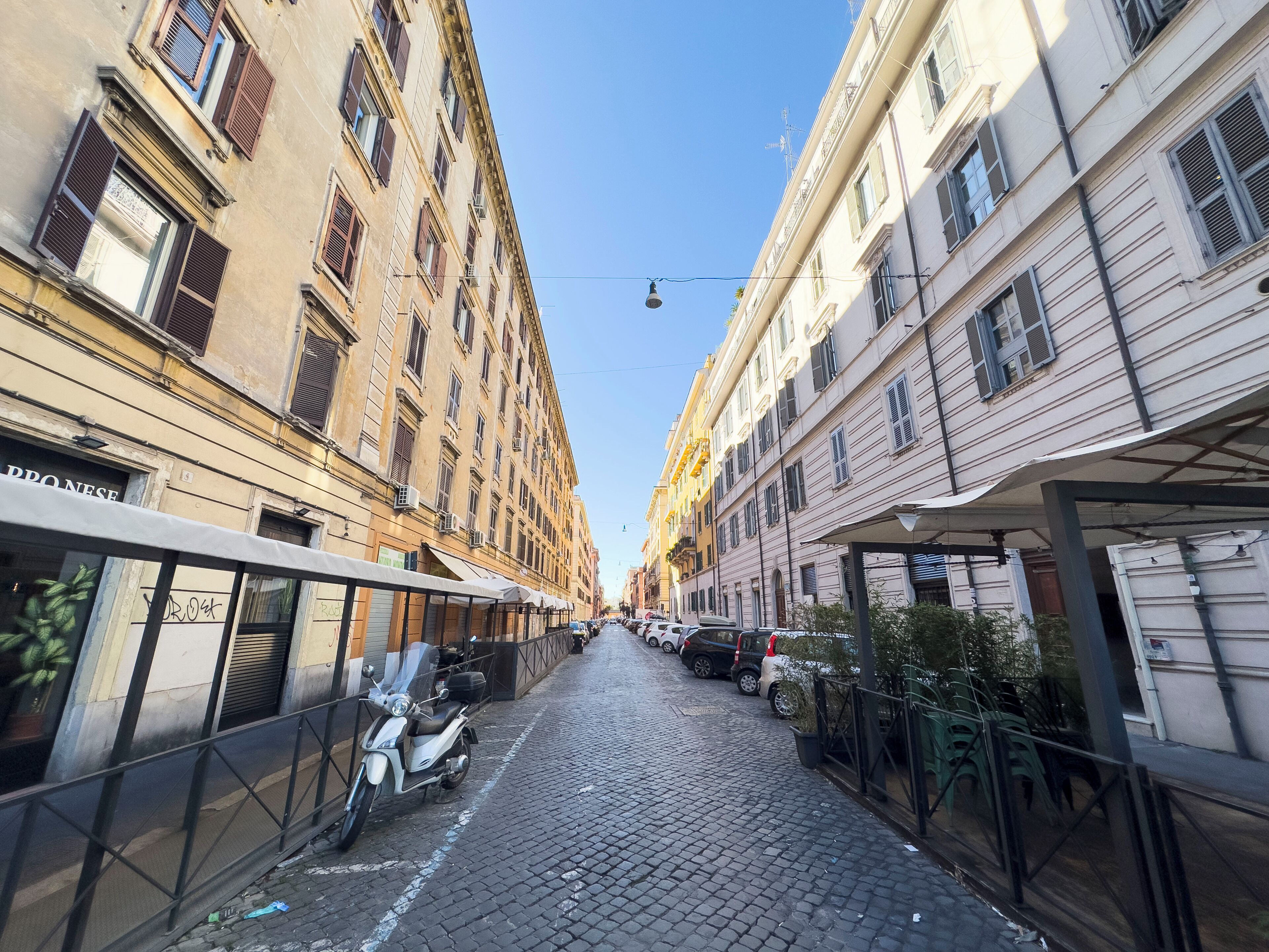 En brostensbelagt gade med historiske bygninger, en parkeret scooter og række af udeservering.