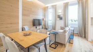 A modern living room with a wooden floor, a dining table with chairs, a sofa, a TV mounted on the wall, and large windows with curtains.