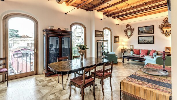 A spacious dining area with a wooden table and chairs, a large window with a view of buildings, and a cozy seating area with a sofa and armchairs. - Trastevere Outstanding 4 Br Terraced Apartment (Rome)