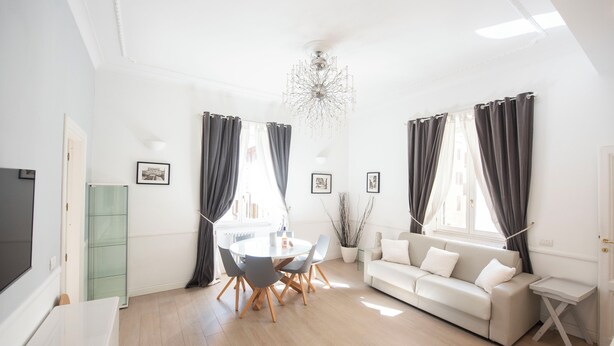 A bright living room with a round dining table, grey chairs, a white sofa, and a chandelier. - Vatican Beautiful 2Br Apartment  (Rome)