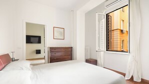 Une chambre à coucher avec un grand lit, une commode, un téléviseur et une fenêtre donnant sur les bâtiments environnants.