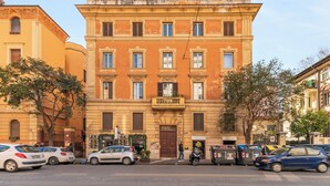 Un immeuble à plusieurs étages doté d’une entrée centrale, de nombreuses fenêtres et d’un petit balcon. On peut voir des voitures et une moto garées devant.