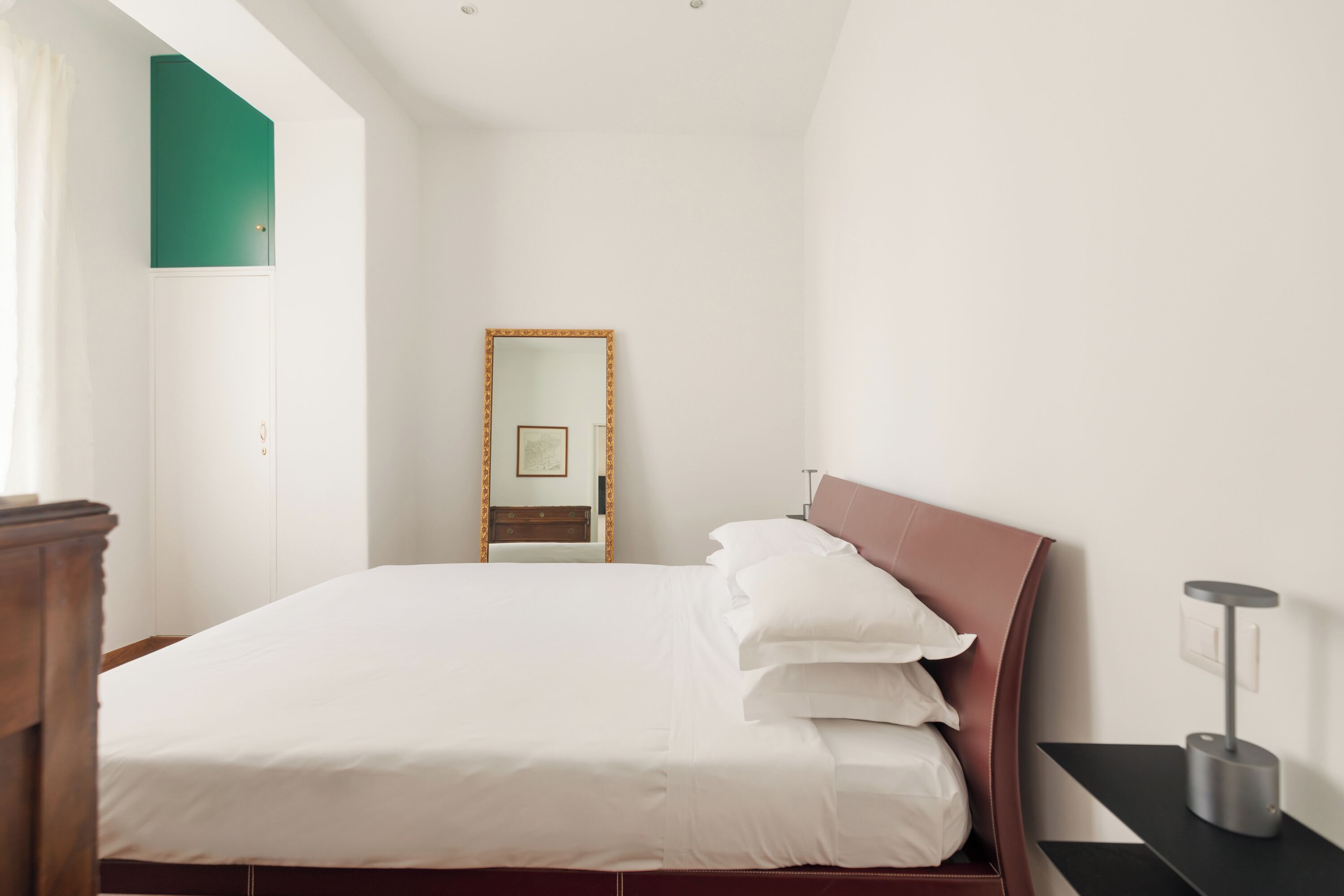 A bedroom with a bed, a bedside table, a mirror, and a wall-mounted shelf.