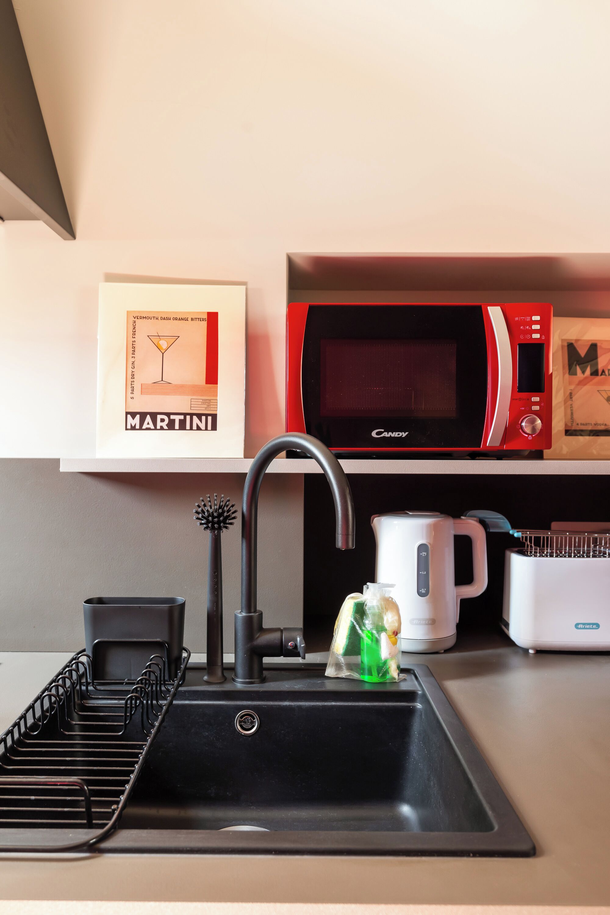 Una cocina con un lavamanos negro, un microondas rojo, una cafetera blanca y un cuadro con un anuncio de Martini.