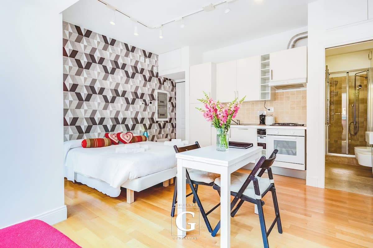 A modern bedroom with a geometric-patterned wall, a white bed, and a small dining area with a table and chairs.