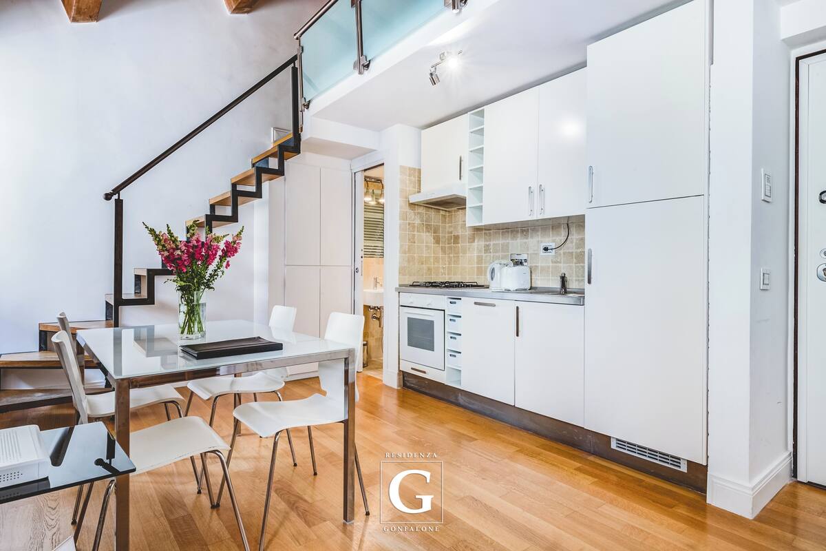 A modern kitchen with white cabinets, a dining area with a glass table, and a staircase.