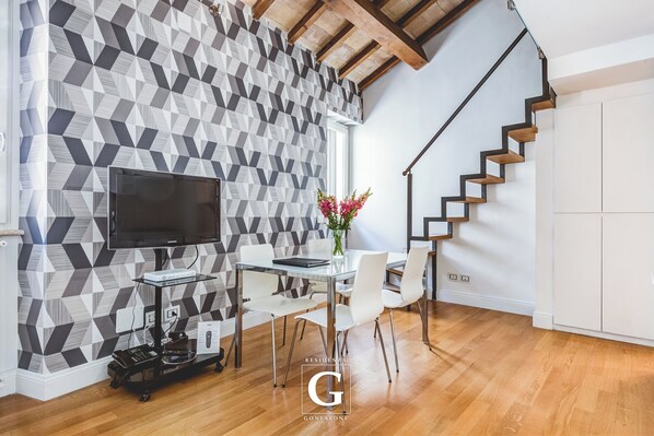 Een moderne woonkamer met een wand met geometrisch patroon, een flatscreen televisie, een glazen salontafel, witte stoelen en een houten trap.