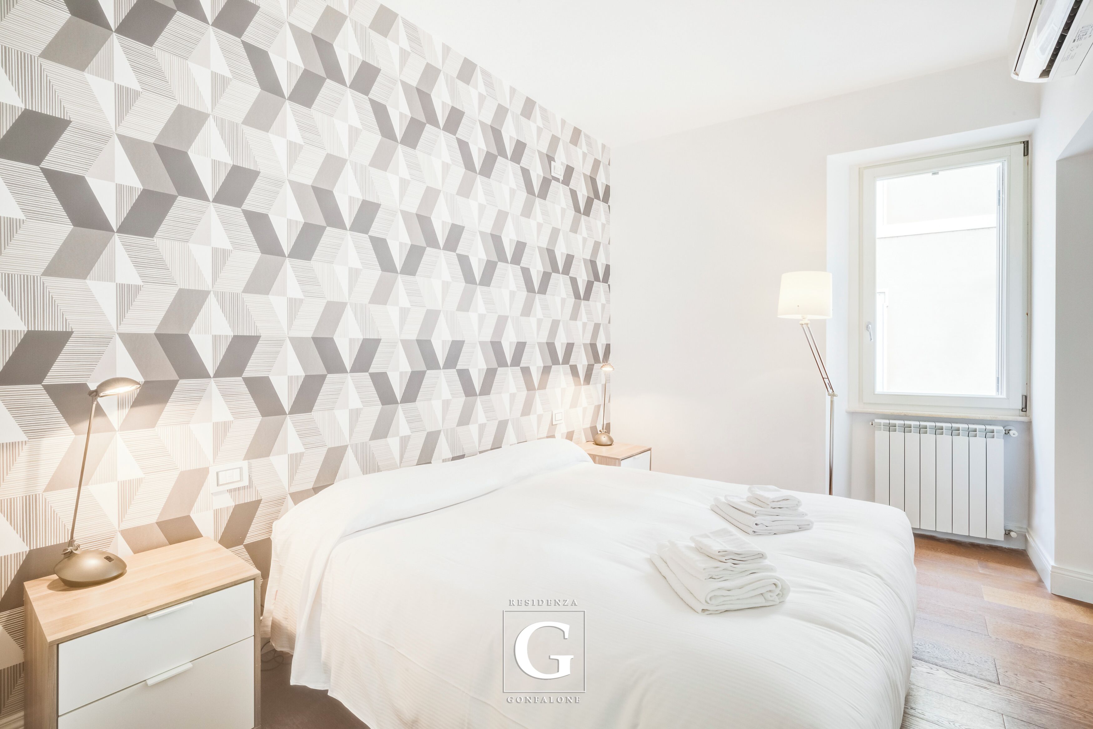 Een moderne hotelkamer met een hoofdeinde met een geometrisch patroon, een netjes opgemaakt bed met gevouwen handdoeken, een bedlamp en een raam met uitzicht.