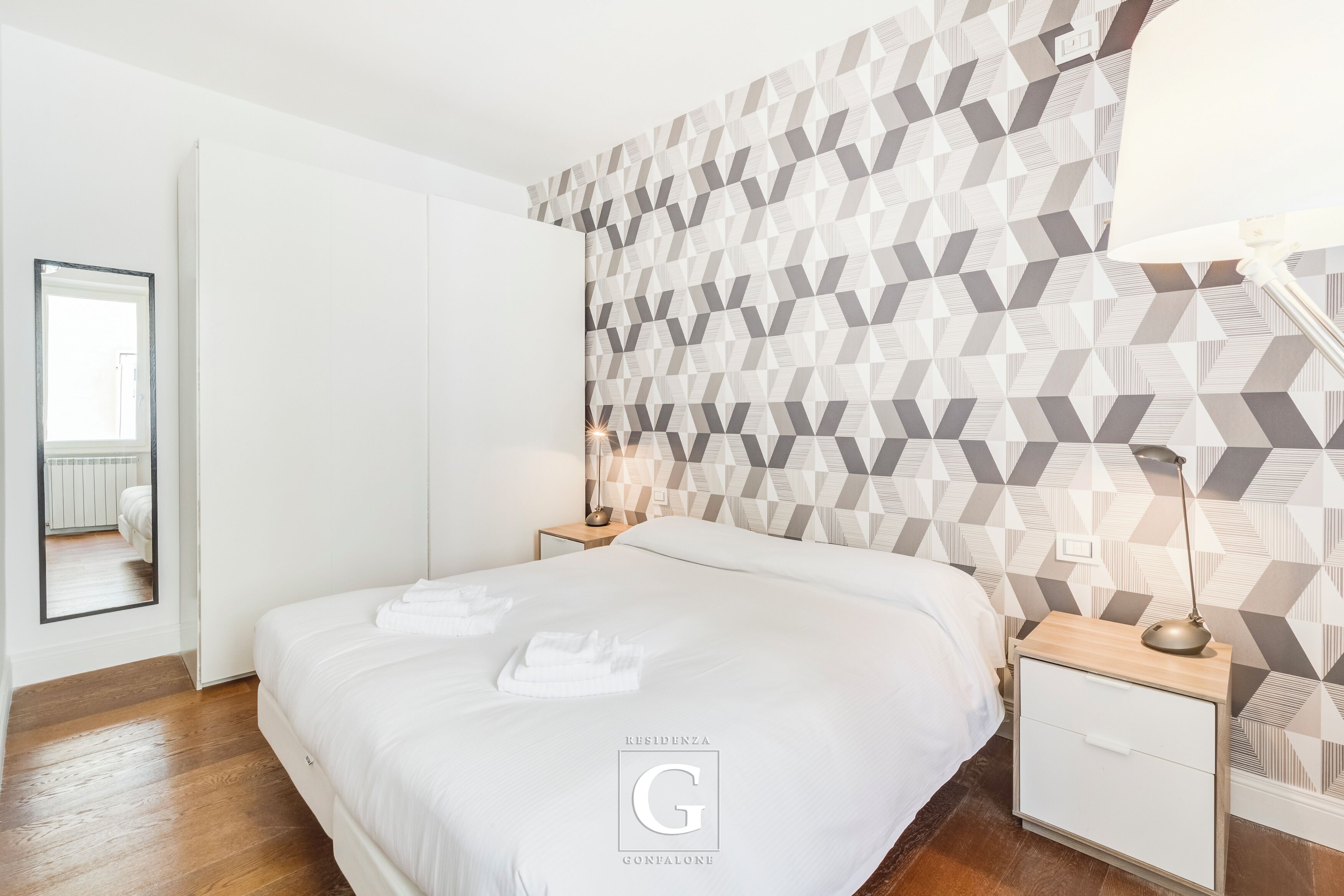 Una habitación de hotel moderna con papel tapiz de estampado geométrico, una cama bien hecha con sábanas blancas, una lámpara de noche y una mesita de noche de madera.