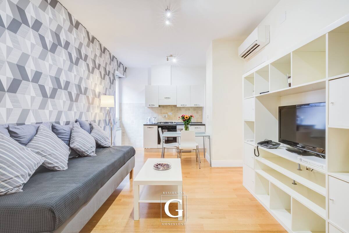 A modern living room with a sofa, a coffee table, and a TV on a stand. There is a kitchen area with cabinets and a dining table.