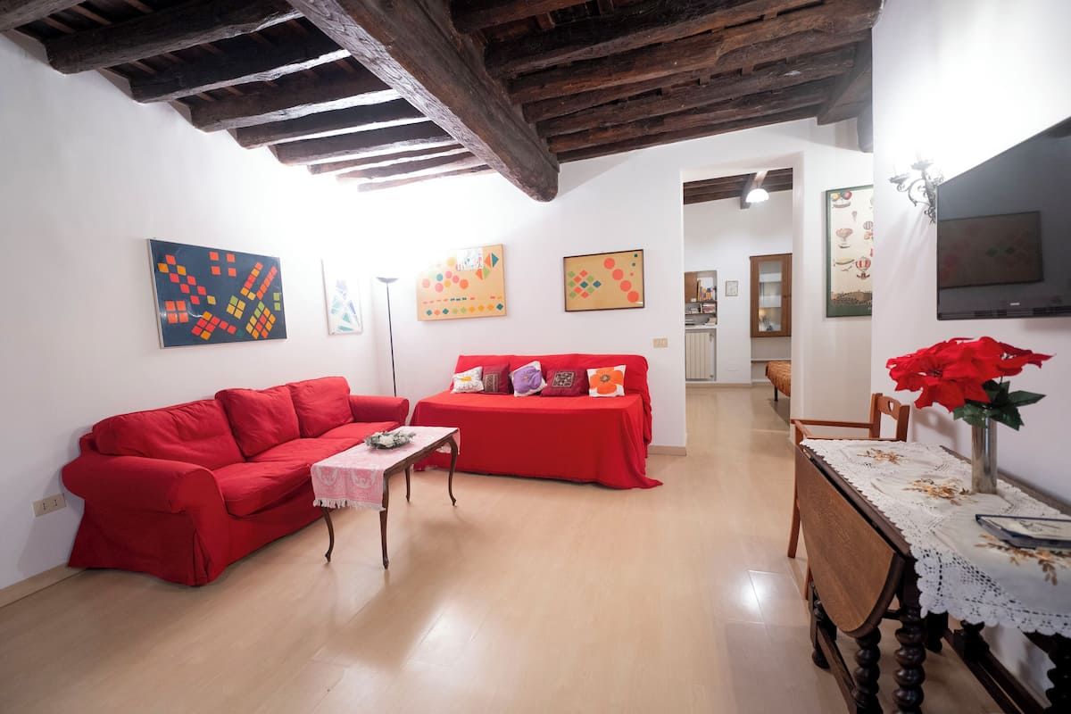 A living room with a red sofa, a small table, and a dining table with chairs. There are paintings on the walls and a TV on the wall.