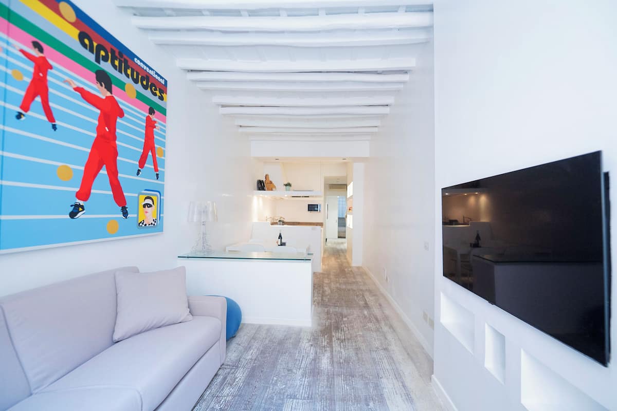 A modern living room with a white sofa, a glass coffee table, a TV mounted on the wall, and a colorful artwork depicting dancers.