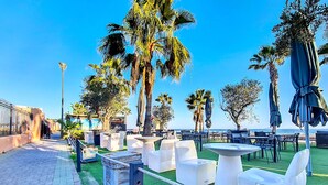 Un espace extérieur aménagé avec des tables et des chaises blanches, entouré de palmiers et offrant une vue sur la mer.
