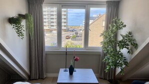 Une chambre moderne avec une grande fenêtre, une petite table ornée d’un vase et d’une bougie, et une plante en pot.