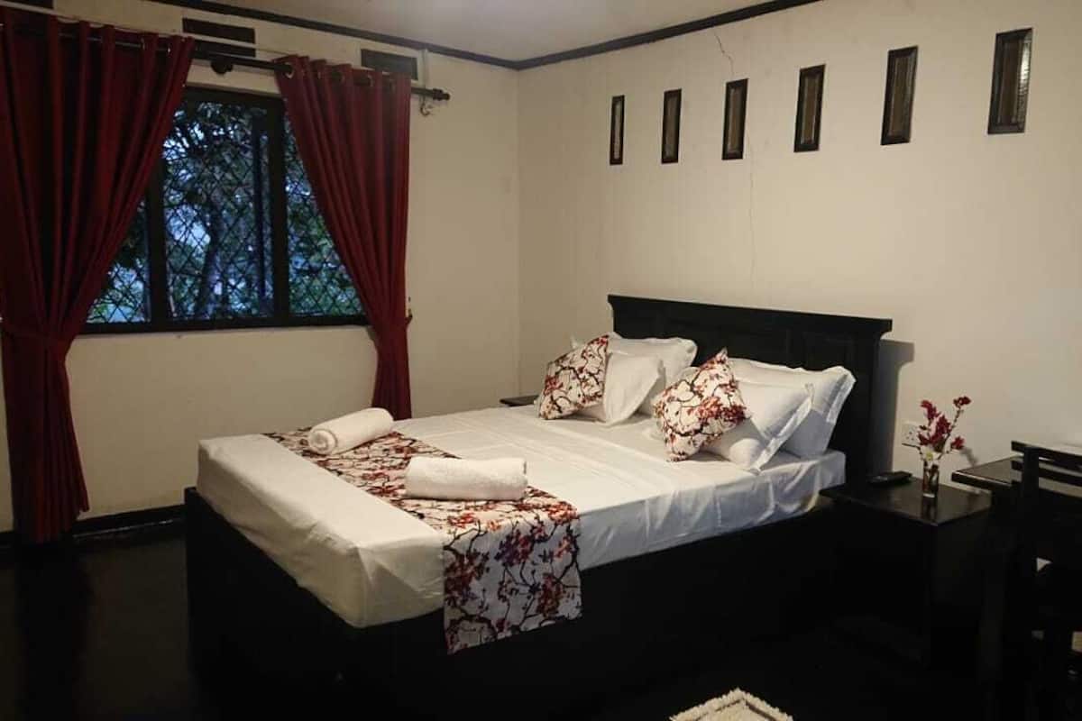 A neatly made bed with white linens and floral-patterned pillows, a bedside table with a vase of flowers, and a window with red curtains.