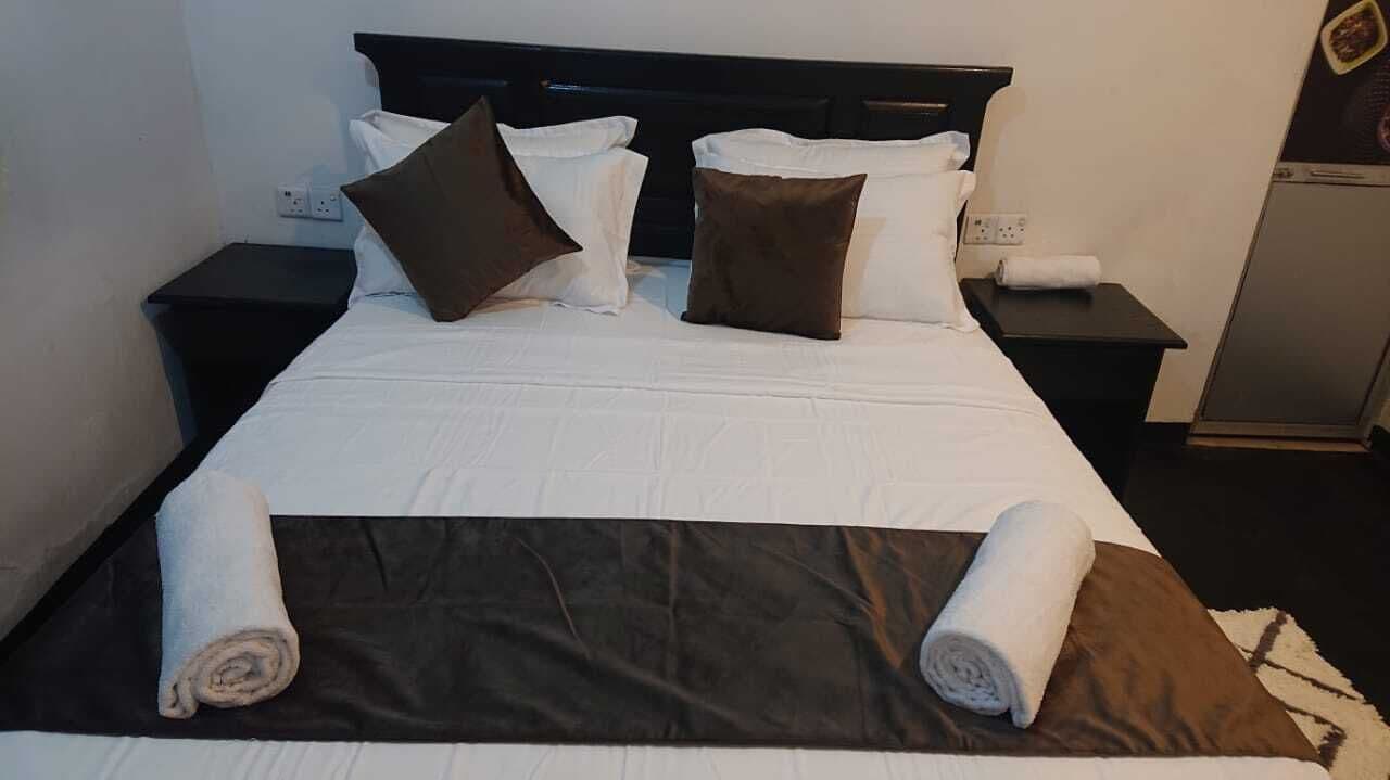 Una cama bien tendida con ropa de cama blanca y marrón, dos mesitas de noche negras y una pequeña mesa con una toalla enrollada a cada lado.