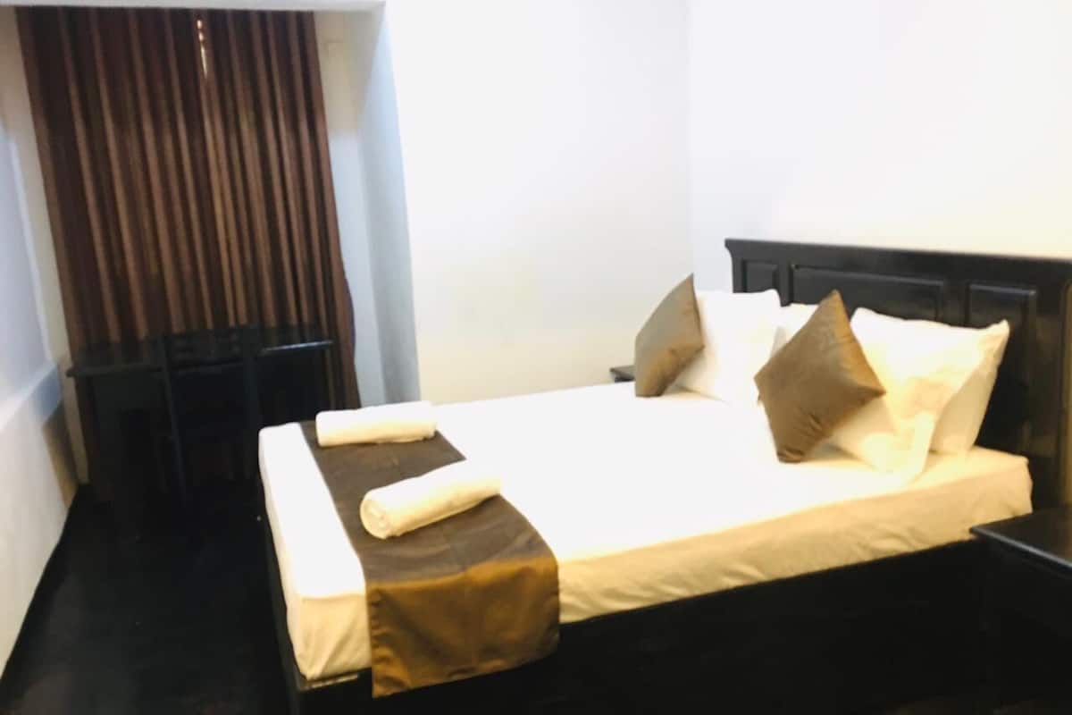 A neatly made bed with a brown bedspread and two pillows, a small bedside table, and a window with vertical blinds.