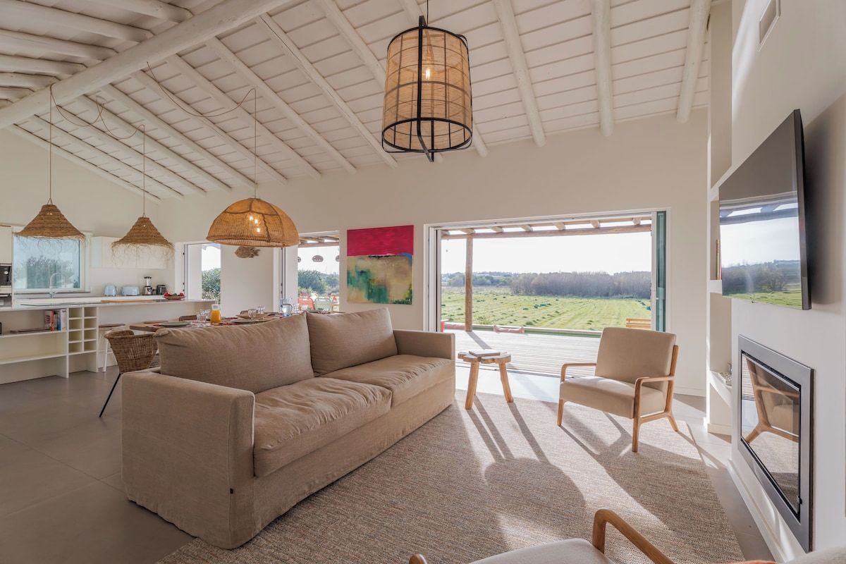Uma sala de estar ampla com sofá, cadeiras, uma mesa de jantar e uma grande janela com vista para o exterior.