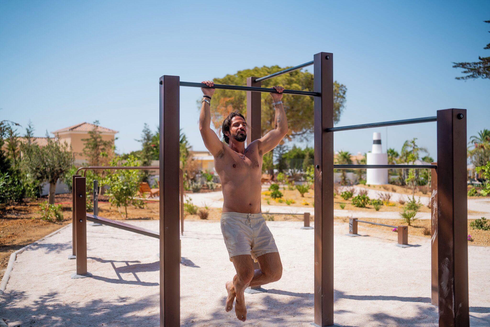 A man performing pull-ups on outdoor gym equipment in a park with trees and buildings in the background.