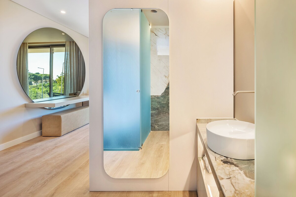 A modern bathroom with a large mirror, a round sink, and a view of greenery outside.