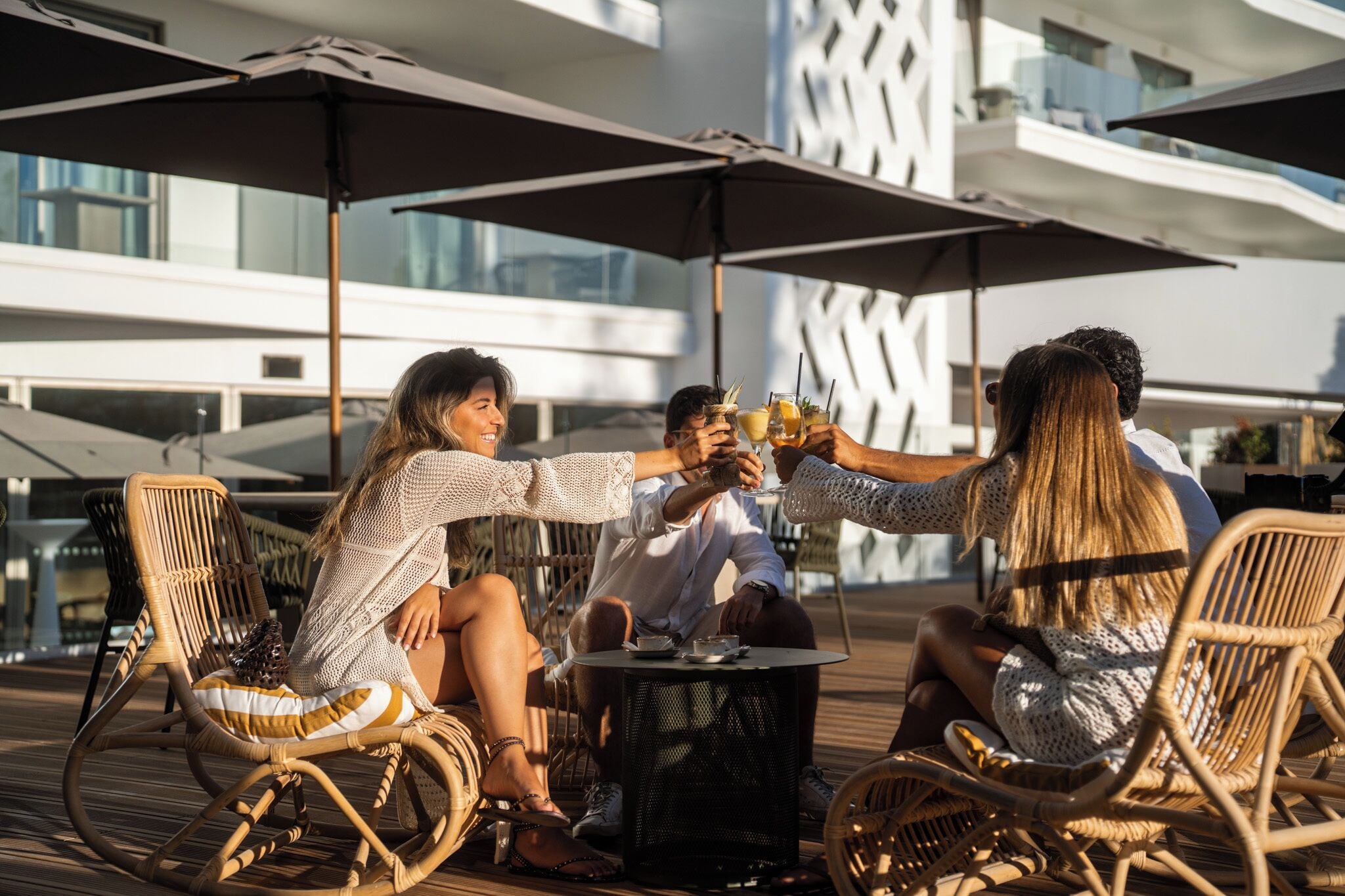 Un grupo de personas brindando con bebidas en una terraza de madera con sillas de mimbre y una mesa redonda, con vista a un edificio moderno con balcones.