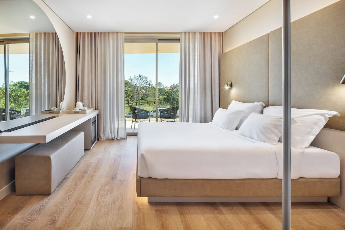 A modern hotel room with a large bed, a desk, and a view of a garden through the window.