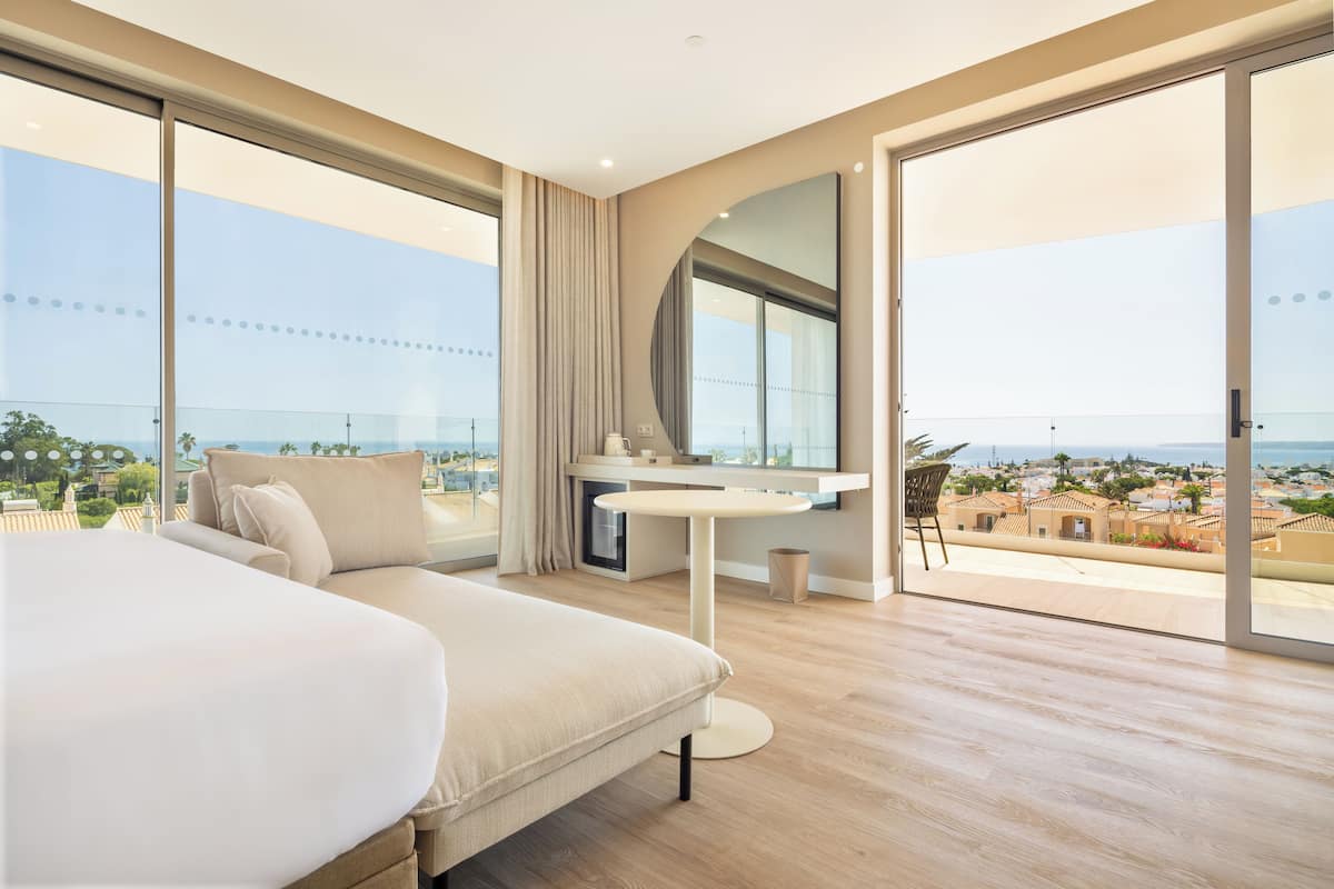 A modern hotel room with a large bed, a round table, and a view of the city and sea.