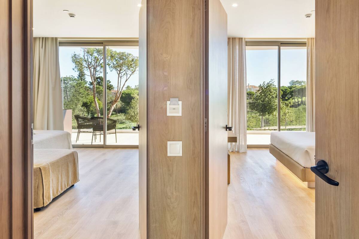 A modern hotel room with a large sliding glass door leading to an outdoor seating area. The room features a wooden floor, a bed, and a small table with a chair.