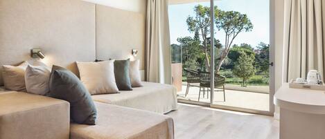 A modern hotel room with a large sliding glass door leading to a balcony with a view of trees and a distant landscape. The room features a sofa set, a small table, and a coffee machine.