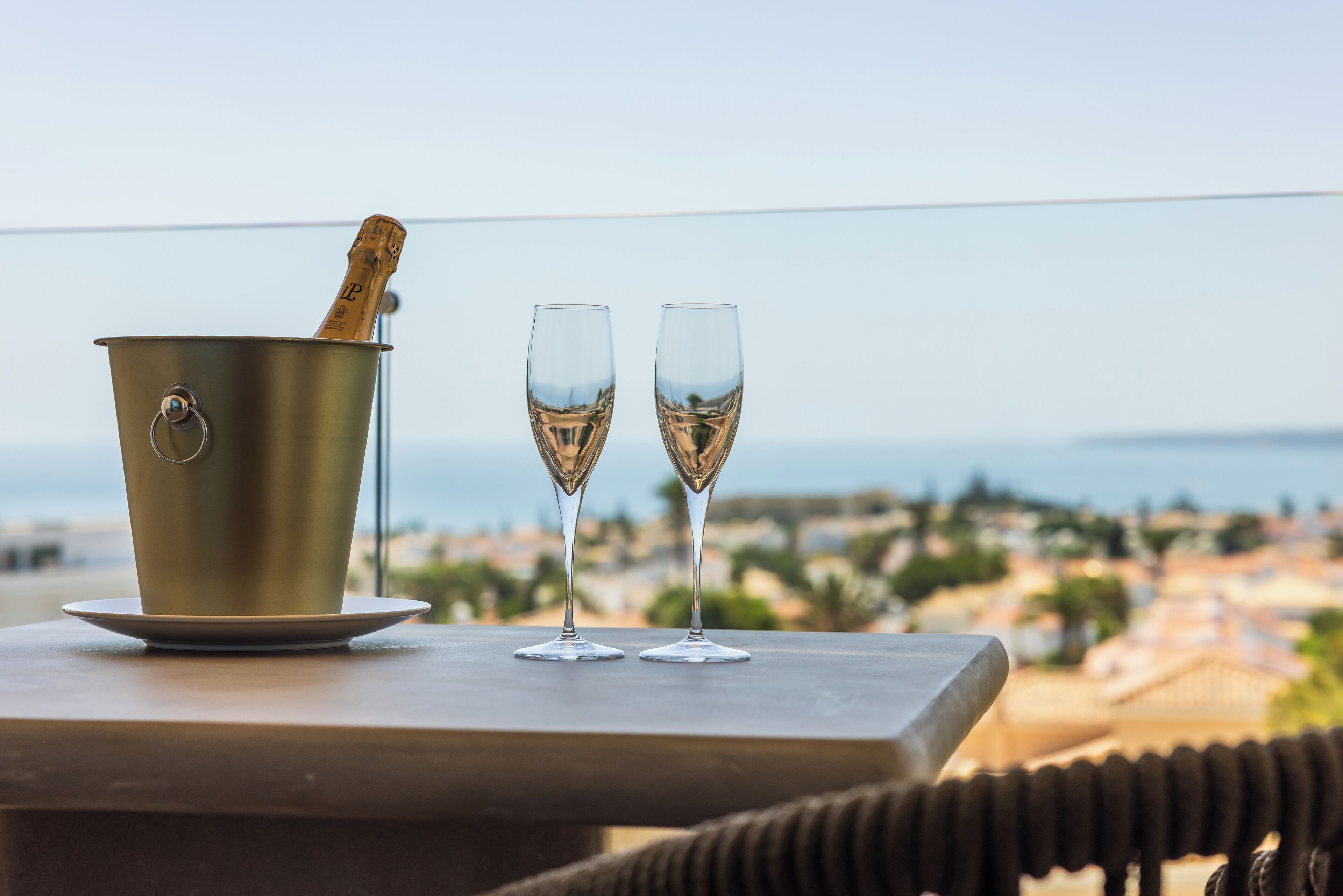 A metal bucket with a cork and two champagne flutes on a table with a view of a coastal town.