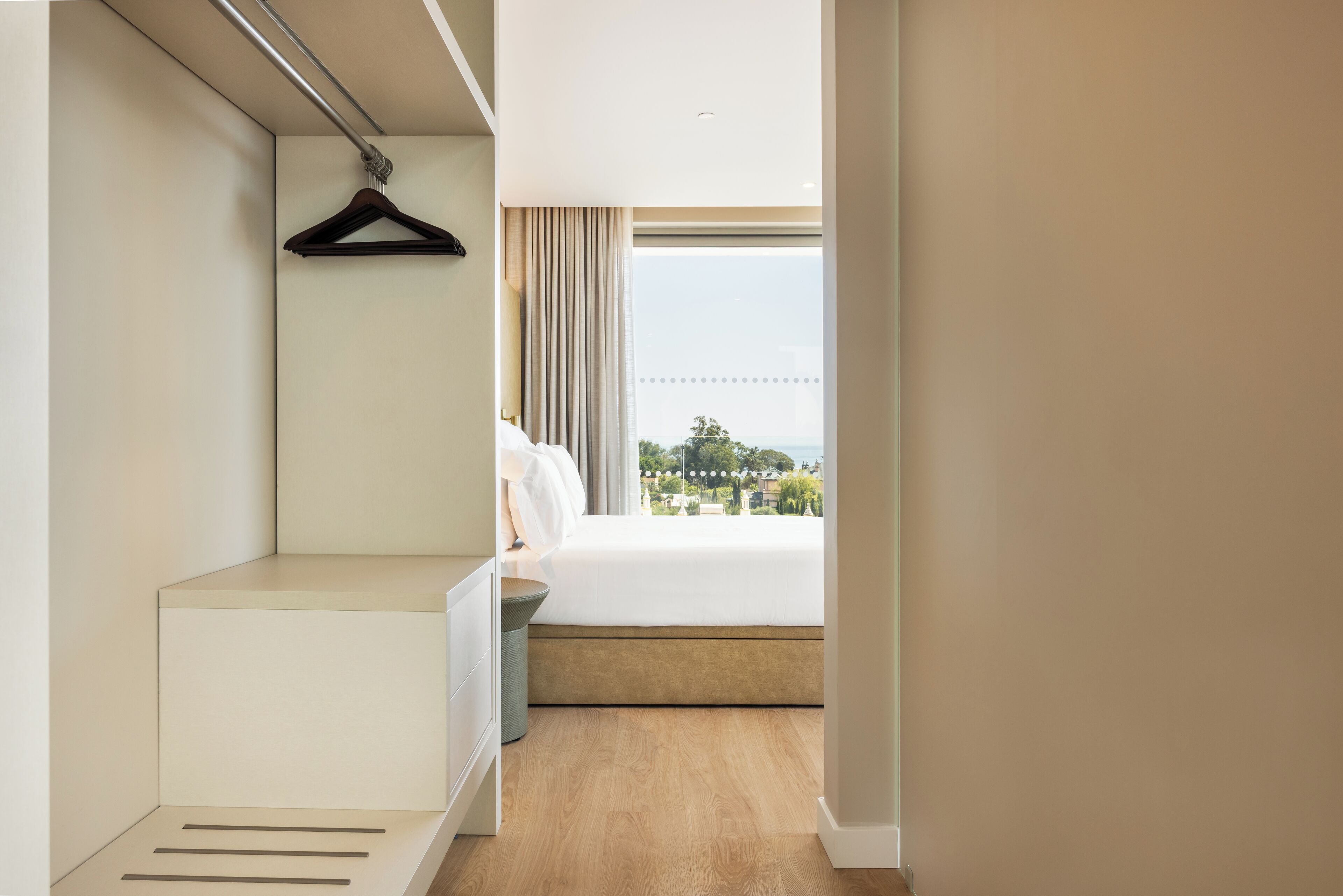 Ein Schlafzimmer mit Bett, Nachttisch, Kleiderhaken und einem Fenster mit Blick ins Freie.
