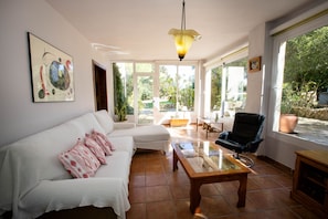 Un salon comprenant un canapé, une table basse et un fauteuil. Un tableau est accroché au mur, et une fenêtre offre une vue sur la verdure à l’extérieur.