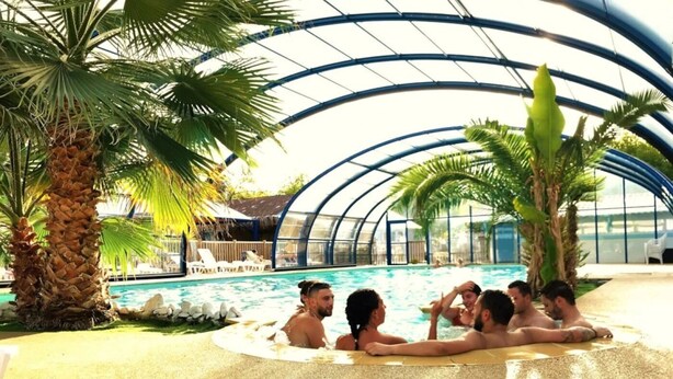 Ein überdachter Innenpool mit Glasdach, Palmen und Liegestühlen.