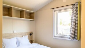 Ein ordentlich bezogenes Bett mit weißen Leinen, ein Buch und ein Hut darauf. Ein Fenster mit gestreifter Vorhang und Blick nach draußen.