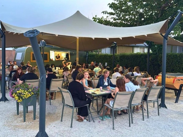 Un espacio exterior para comer con mesas y sillas, un área de descanso techada y un edificio al fondo.