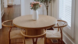 Un comedor con una mesa redonda de madera, cuatro sillas de mimbre y un jarrón blanco con flores.
