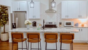 Una cocina moderna con armarios blancos, un refrigerador de acero inoxidable, horno empotrado, microondas y un lavamanos. Hay tres taburetes de madera y una planta en maceta.