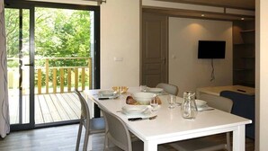 Un coin repas avec une table blanche dressée pour quatre personnes, une porte-fenêtre coulissante donnant sur une terrasse extérieure et un téléviseur à écran plat fixé au mur.