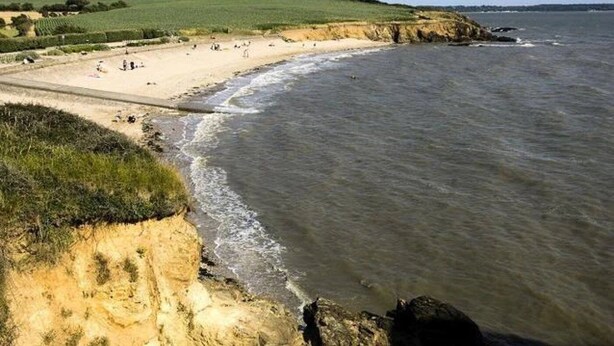 A coastal beach with a sandy shore, grassy dunes, and a rocky outcrop.