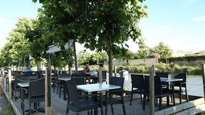 Un área de comedor al aire libre con mesas y sillas, rodeada de follaje y una masa de agua.