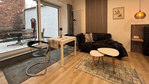 A modern living room with a black sofa, a small table, and a coffee table. There is a large window with a view of the outside, a radiator, and a wall-mounted air conditioner.