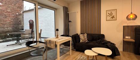 A modern living room with a black sofa, a small table, and a coffee table. There is a large window with a view of the outside, a radiator, and a wall-mounted air conditioner.