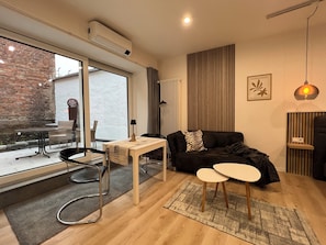En moderne stue med en sort sofa, et lille bord og et sofabord. Der er et stort vindue med udsigt udendørs, en radiator og en vægmonteret aircondition.