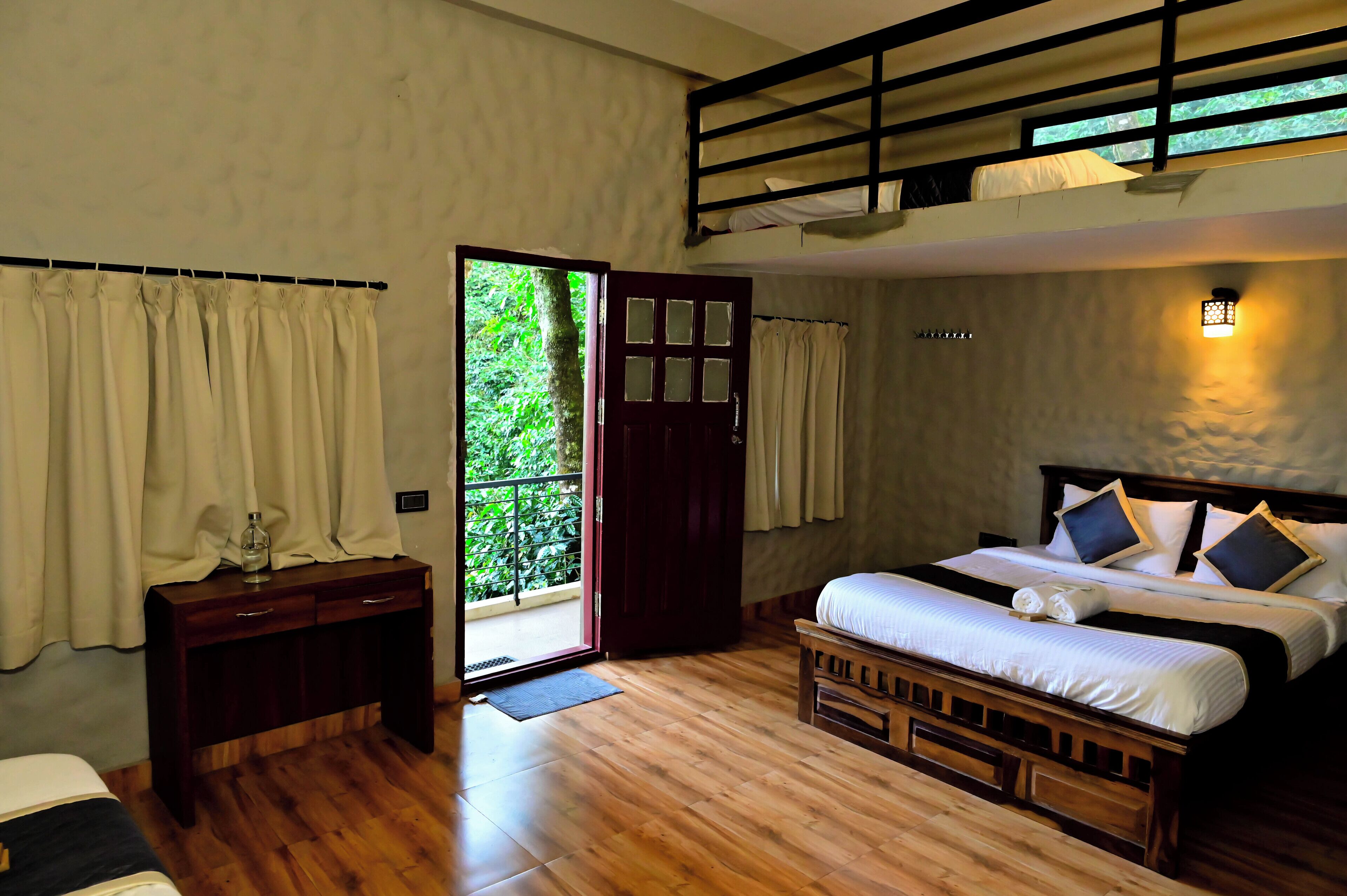 Une chambre avec un plancher en bois, un lit avec une literie blanche, un bureau avec des tiroirs, une fenêtre avec des rideaux et une porte-fenêtre donnant sur un balcon.