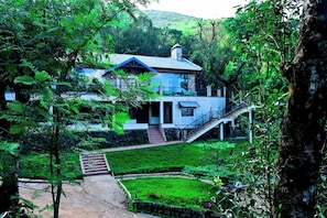 A house with a staircase leading to a balcony, surrounded by lush greenery and a well-maintained lawn.