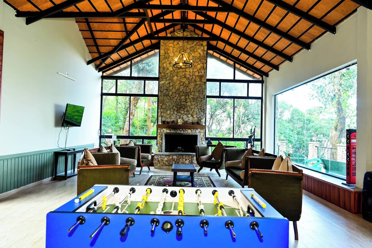 A room with a foosball table, comfortable seating, a stone fireplace, and large windows offering a view of greenery.
