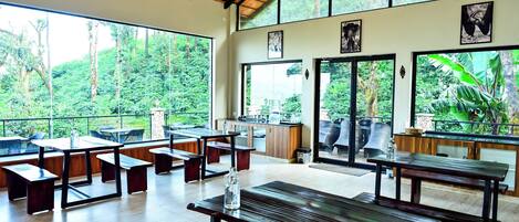 A spacious indoor area with wooden tables and benches, large windows offering views of a lush forest.
